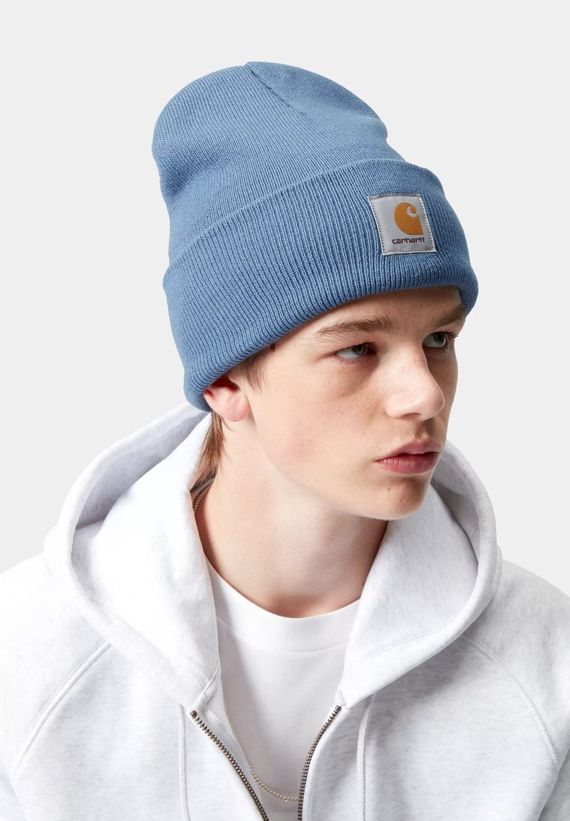 Joven con un gorro de lana azul claro con el logo de Carhartt, sudadera blanca con cremallera y camiseta blanca, mirando ligeramente hacia la derecha.