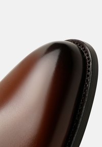 Chaussure en cuir marron avec une finition polie, bout rond et couture contrastante le long du bord. Texture lisse et éclat subtil.