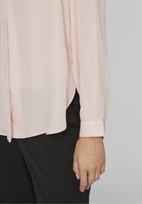 Blusa rosa chiaro a maniche lunghe con tessuto liscio e leggermente trasparente. Presenta un orlo curvo e polsini con bottoni, indossata con pantaloni neri.