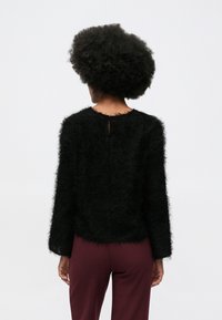 Afro frizura viselője fekete bolyhos hosszúujjú felsőben és bordó nadrágban, hátulról látható, fehér háttér előtt.