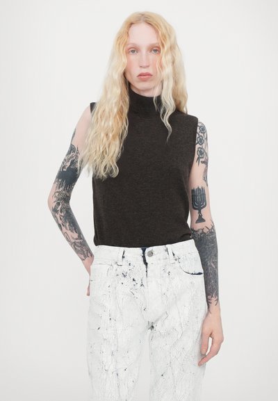 Sleeveless turtleneck in dark brown fabric paired with white jeans featuring a cracked pattern and blue accents. Intricate tattoos visible on arms.