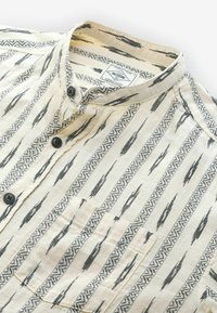 Chemise à manches courtes couleur crème avec des motifs géométriques noirs. Dotée d'un col à rabat et d'une poche poitrine avec fermeture à bouton.