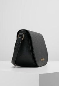 Svart läderhandväska med en texturerad yta, prydd med en guldlogo och ett lockstängningssystem, placerad på en vit yta med en enkel form.