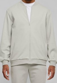 Ljust grå ziptröja med rund hals, ribbade ärmslut och nederkant, tillverkad av mjukt tyg, bärs över en vit T-shirt.