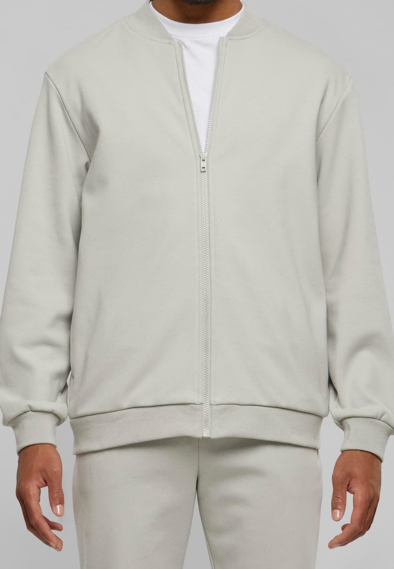 Ljust grå ziptröja med rund hals, ribbade ärmslut och nederkant, tillverkad av mjukt tyg, bärs över en vit T-shirt.