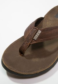 Brune lærflip-flop med en teksturert stropp og en polstret såle. Har en gummisåle og en liten metalllogo på stroppen.