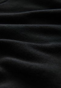 Juodas audinys su minkštais raukšlėmis ir matoma mezgimo tekstūra pritemdytoje šviesoje, pabrėžiant sklandumą ir medžiagos detalumą.