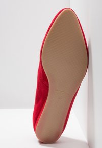 Chaussure plate rouge avec semelle beige texturée et bout pointu, positionnée verticalement contre un coin blanc entre le mur et le sol.
