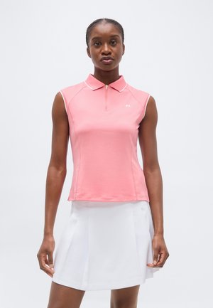 Kobieta ubrana w różową, bezrękawną koszulkę polo z kołnierzykiem i białą plisowaną spódnicę, stojąca na tle jednolitego jasnego tła.