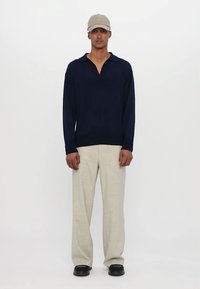 Polo a maniche lunghe color navy con colletto rilassato, abbinato a pantaloni in lino di colore chiaro e dalla vestibilità ampia e scarpe nere.