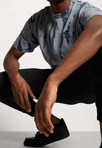 Nike tričková košile s tie-dye vzorem v šedé barvě s námořnickými modrými pruhy, v kombinaci s černými kalhotami a černými teniskami, která se vyznačuje uvolněným střihem a jednoduchým designem.