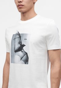 Homme portant un t-shirt blanc avec un motif chauve-souris noir et gris et le mot BOSS sur la poitrine.