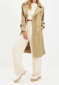 Béžový trenčkot s dvojitým zapínáním, velkým límcem a knoflíkovými detaily, doplněný světlými širokými kalhotami a teniskami na platformě.