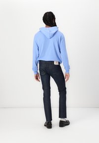 Personne avec des cheveux tressés portant un sweat à capuche bleu, un jean sombre, des chaussettes blanches et des chaussures noires, se tenant contre un fond blanc, de dos.