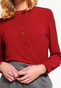 Blouse rouge à manches longues avec un col rond, un devant boutonné et des boutons noirs. Tissu texturé avec une finition lisse. Jupe grise partiellement visible.