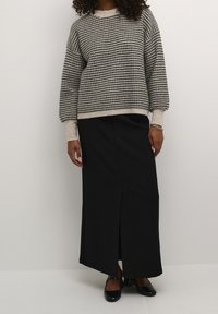 Beige und schwarz gemusterter Pullover in lockerer Passform, kombiniert mit einem langen schwarzen Rock mit seitlichem Schlitz und schwarzen Schuhen.