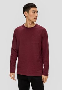 Bordowa koszula z długim rękawem, uszyta z miękkiego materiału, z okrągłym dekoltem i kieszonką na lewej piersi, w zestawie z czarnymi spodniami.