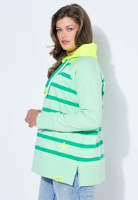Mujer con una sudadera con capucha a rayas verde menta y capucha de color amarillo neón, de perfil contra un fondo blanco.