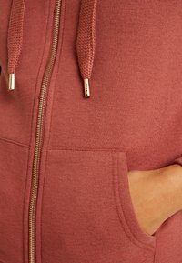 Gros plan d'un sweat à capuche zippé couleur rouille avec une fermeture éclair dorée et des embouts de cordon, montrant la main d'une personne dans la poche avant.