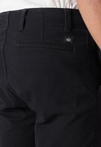 Black cotton trousers featuring a smooth texture, a rear pocket with a flap, and a small logo tag. The waistband is neatly stitched.