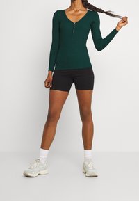 Grünes, geripptes Langarmshirt mit Reißverschluss am Kragen, kombiniert mit schwarzen Shorts und leichten Sneakers, zeigt ein figurbetontes Design und strukturstoff.