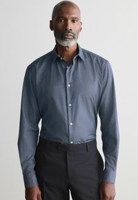 Camisa de vestir azul con estampado, con cuello, mangas largas y parte delantera abotonada. Combinada con pantalones oscuros y una textura suave.