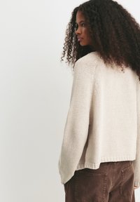 Femme avec de longs cheveux bouclés, portant un pull en tricot crème et un pantalon marron, debout et regardant sur le côté devant un fond uni.