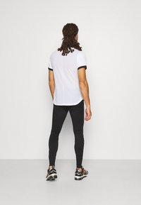 Weißes sportliches T-Shirt mit schwarzem Saum, kombiniert mit schwarzen, eng anliegenden Leggings und Turnschuhen. Sichtbare Nähdetails auf dem Shirt.