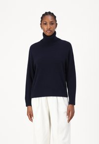 Pulover cu guler înalt de culoare bleumarin, cu o croială lejeră, manșete și tiv cu nervuri, și fante laterale. Împerecheat cu pantaloni de culoare deschisă, lejeri.