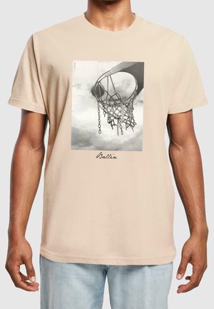 Man die een beige T-shirt draagt met een zwart-witte foto van een basketbalring en net, met het woord "Ballin" onder de afbeelding geschreven.