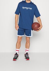 Marineblauer Sporthoodie mit dem Text "Breakfast Club", luftige Basketballshorts mit weißem Saum, schwarz-rote Sneakers mit roten Akzenten und rot-gestreifte weiße Socken.
