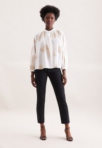 Blusa bianca con motivo floreale beige, scollatura arricciata e maniche lunghe, abbinata a jeans neri aderenti e tacchi a spillo neri.