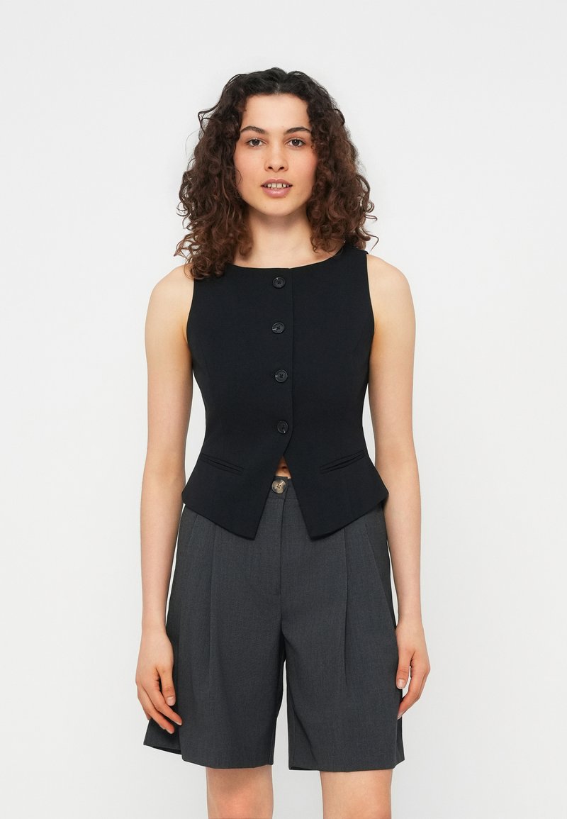 Γυναίκα με σγουρά μαλλιά που φοράει αμάνικο μαύρο γιλέκο με κουμπιά και γκρι ψηλόμεσα σορτς με πιέτες, όρθια μπροστά σε απλό λευκό φόντο.