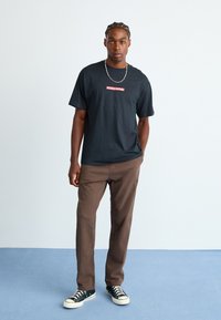 Homme aux cheveux tressés portant un t-shirt noir Quiksilver, un pantalon marron, des baskets noires et un collier en chaîne argenté, sur un sol bleu devant un mur blanc.