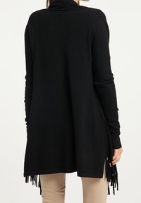 Gilet en tricot noir avec un col haut, des manches longues et des fentes sur les côtés, présentant des détails en franges au niveau de l'ourlet. Coupe décontractée et confortable.