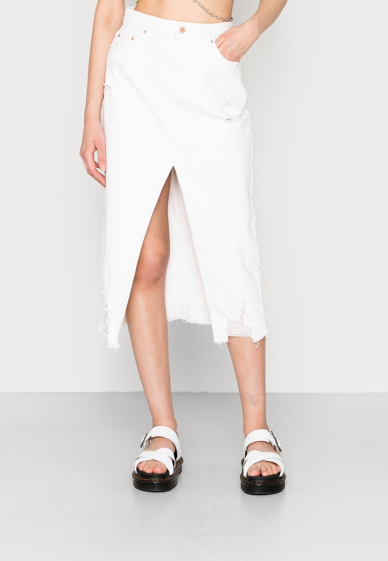 Jupe midi en denim blanc avec ourlet effiloché et fente devant. Associée à des sandales blanches avec des lanières larges et une semelle noire épaisse.
