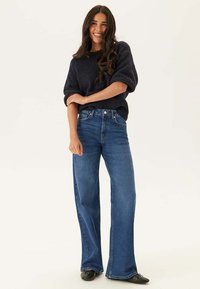 Mörkblå stickad tröja med korta, voluminösa ärmar, kombinerad med högmidjade, vida blå jeans. Modellen står mot en ljus bakgrund.
