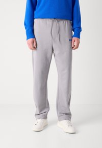 Pantalones grises con cintura elástica con cordón, textura suave, bolsillos laterales y un pequeño logo azul en el muslo izquierdo. Combinados con un suéter azul.