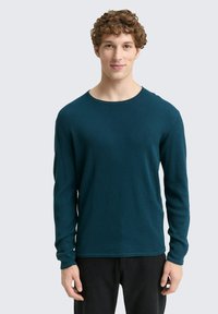 Cyaan lange mouwen shirt met een gestructureerd patroon en ronde halslijn, gecombineerd met donkere broeken. De stof lijkt zacht en rekbaar.