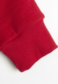 Cuff di un maglione rosso con texture a costine, tessuto morbido a maglia e una leggera svasatura al bordo. Finitura liscia senza abbellimenti visibili.