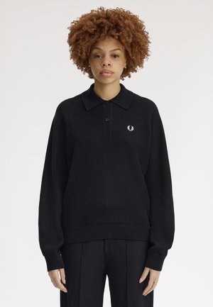 Femme aux cheveux bouclés portant un pull en maille noir à manches longues avec un petit logo blanc sur la poitrine, debout devant un fond uni.