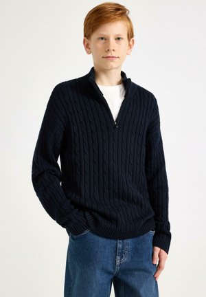 Maglione blu navy a maglia grossa con collo con cerniera, polsini e orlo a coste. Abbinato a jeans azzurri chiari. T-shirt bianca tinta unita sotto.