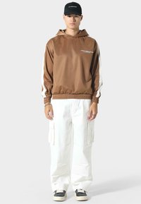 Bruine hoodie met witte zijpanelen, elastische manchetten en tailleband. Gepaard met losse witte cargo-broeken en zwarte sneakers.