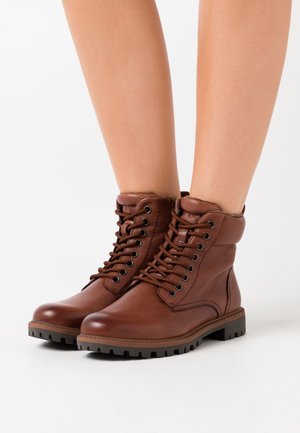 Braune Leder-Stiefel bis zum Knöchel mit einer robusten Sohle, Schnürverschluss, gepolstertem Schaft und strukturierter Oberfläche. Mit schwarzen Metallösen und Nähten.