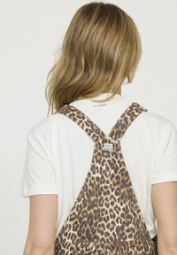 Salopette en imprimé léopard avec bretelles ajustables, fabriquée en denim. T-shirt blanc partiellement visible en dessous. Étiquette de marque sur la bretelle.