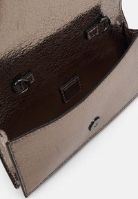 Kiiltävä tummanruskea nahkakäsilaukku, jossa on teksturoitu pinta, avoin läppä, metallinen solki ja näkyvä tuotemerkin etiketti sisällä.