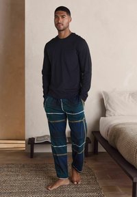 Mies seisoo paljain jaloin makuuhuoneessa musta pitkähihainen paita ja vihreät ruudulliset pyjamahousut päällään, kudotun maton päällä lähellä sänkyä.