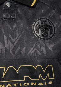 Crna polo majica s teksturiranim geometrijskim uzorcima, crnim kružnim logotipom i istaknutim žutim imenom branda otisnutim velikim slovima.