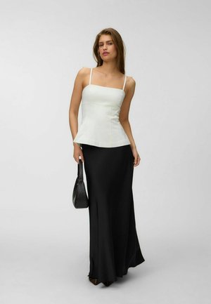 Femme portant un haut blanc sans manches et une longue jupe noire, tenant un petit sac à main noir, debout devant un fond clair uni.