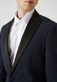 Giacca da smoking blu navy con rever in raso nero, taschino sul petto, e chiusura a bottone; tessuto testurizzato con vestibilità su misura. Camicia bianca da dress visibile sotto.